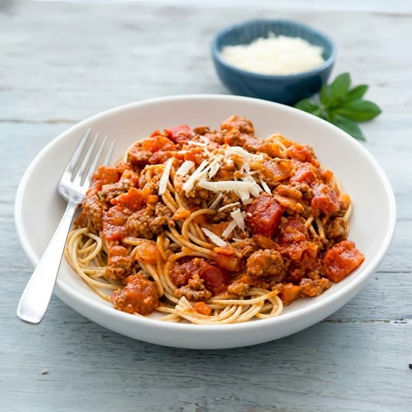 A white bowl of spaghetti with meat sauce and shredded cheese, with a fork, on a wooden table.